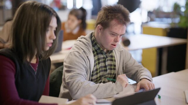 Two young students are collaborating on an assignment at a table in a classroom setting. A female student is helping a male student with Down syndrome with his studies.