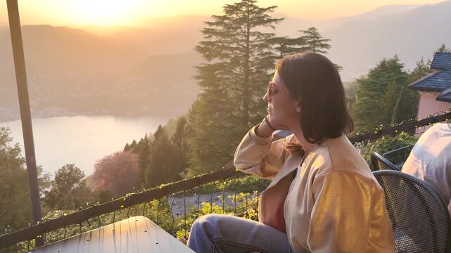 Young woman sitting on terrace in Brunate overlooking Lake Como at sunset, enjoying peaceful scenic mountain view and warm golden hour travel moment in Italy