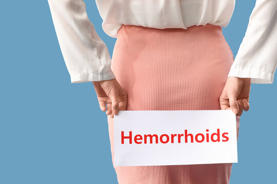 Young woman holding paper with word HEMORRHOIDS on blue background, back view