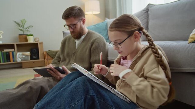 Stab shot of gen Alpha daughter handing homework notebook to Caucasian father giving her digital tablet in exhange as reward sitting on home floor