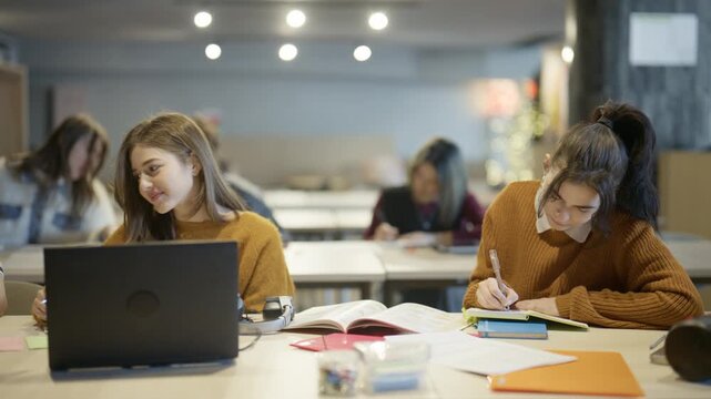 Two young female students concentrate on their studies, writing in notebooks and using a laptop in a modern classroom.