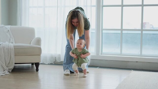 Baby development. First steps. Little infant girl making first steps at home. Happy mother supporting baby first steps. Mom helping toddler child walking in living room. Happy family at home