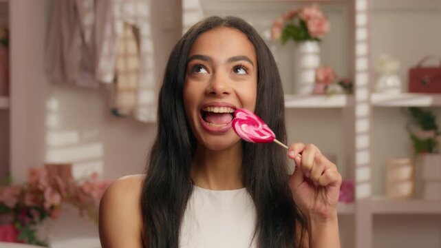 Smiling Hispanic girl biting sweet sugar candy with healthy teeth looking at camera happy Latina 20s woman American female bite lollipop smile in pink bathroom diet nutrition food natural cosmetic
