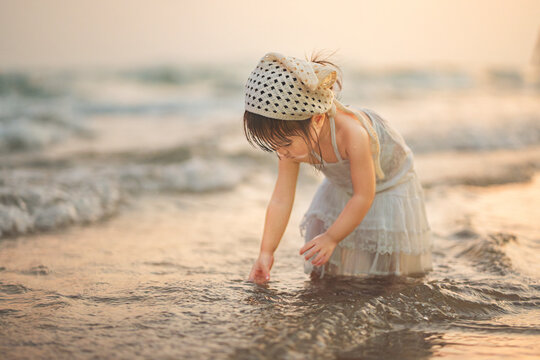 Cute Asian toddler girl playing with sea water on a tropical beach at sunset. Little child wearing a white dress and bandana exploring nature during summer family vacation, reflecting wellness