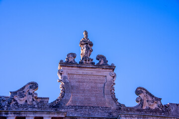 The upper section of Porta San Biagio in Lecce, featuring a statue of Saint Blaise above an inscribed pediment, framed by elegant baroque scrolls against the clear sky. © lucazzitto