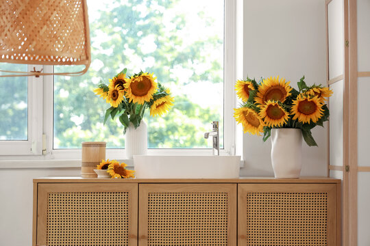 Vases with bouquets of beautiful sunflowers on wooden cabinet in bathroom