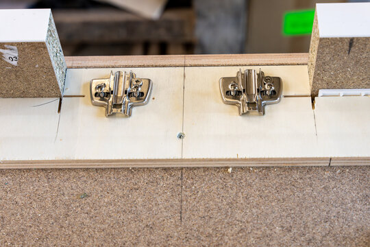 Installation de charni&egrave;res m&eacute;talliques sur un meuble en m&eacute;lamin&eacute; blanc et bois dans un atelier de montage.