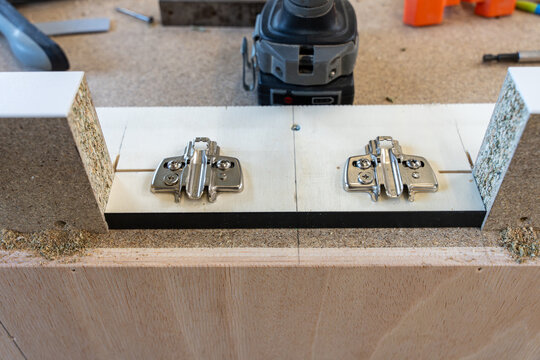 Installation de charni&egrave;res m&eacute;talliques sur un meuble en m&eacute;lamin&eacute; blanc et bois dans un atelier de montage.