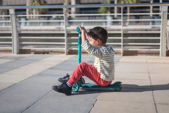 A cheerful five-year-old boy sits on a turquoise scooter, playing on a smooth, paved surface in a vibrant urban environment