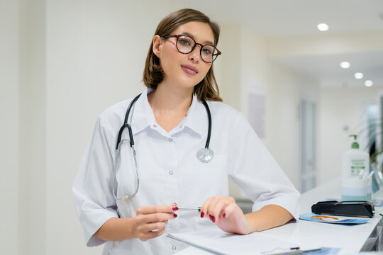 Portrait of young woman doctor standing by reception desk in hospital corridor, confident medical professional in clinic hallway, healthcare, medicine, patient care concept