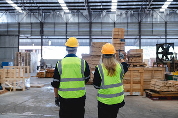 Naklejka na ściany i meble Two Caucasian workers conduct detailed warehouse inspection, pointing at machinery amidst stacked wooden pallets and boxes in a bright, industrial factory setting, wearing high-visibility yellow vests