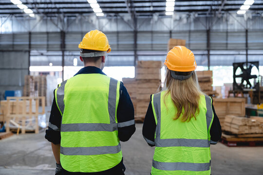 Caucasian workers conduct warehouse inspection amidst stacked boxes and wooden pallets in busy industrial setting with high-visibility vests.
