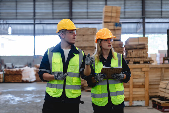Two Caucasian Workers Conduct Detailed Warehouse Inspection with Measuring Tools and Safety Vests in a Cluttered Wooden Factory Environment for Quality Control Assessment.