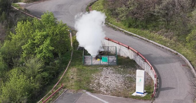 Larderello, Italy - 15 April 2026: Aerial view of Enel Green Power geothermal plant with steam rising from a fenced vent near a winding road and green trees in Tuscany.