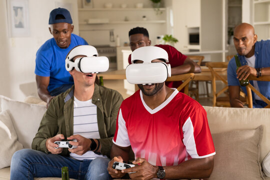 Diverse male friends playing VR on sofa with headsets and controllers, spectators holding bottles
