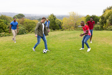 Obraz premium Diverse male friends playing soccer on lawn, dribbling black-and-white ball, wearing green jacket