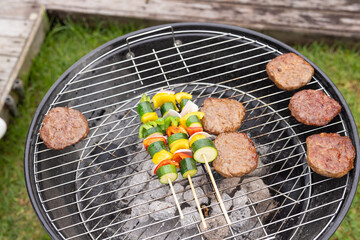 Charcoal kettle grill sitting on lawn near deck, holding six beef patties and three veg skewers © wavebreak3