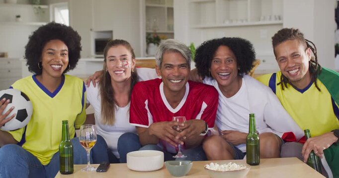 Diverse friends in jerseys on couch reacting to goal on TV, laughing, holding soccer ball, drinks