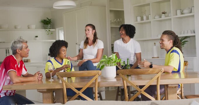 Diverse friends in jerseys at home by table, woman in white tee speaking touching shoulder toasting