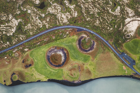 Aerial view of pseudo craters and winding road contrasting against the lush green landscape and the blue waters of Lake Myvatn, Iceland.