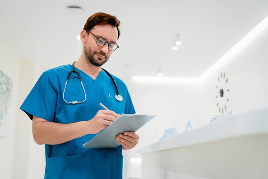 Professional portrait of young male surgeon with clipboard in hospital hallway, confident doctor in medical environment, healthcare, clinic service, patient care and staff concept copy space
