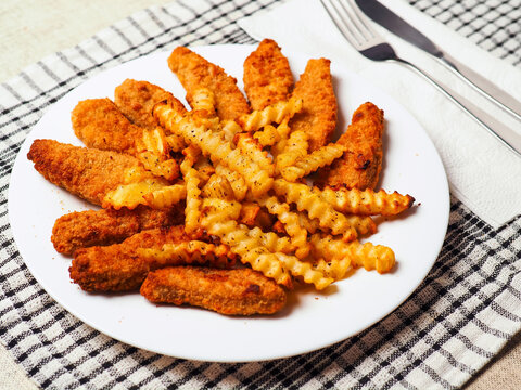 Golden chicken nuggets or goujons sit beside crispy crinkle cut potato fries. Plate rests on checkered cloth with fork and knife nearby. Simple meal suggests comfort and satisfaction