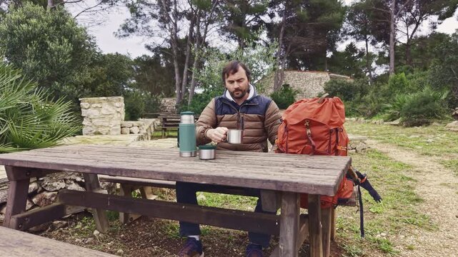 Man resting at picnic table drinking from mug during hiking trip.