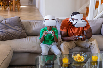 African American father and son playing VR on sofa wearing headsets with controllers, chips, juice © wavebreak3