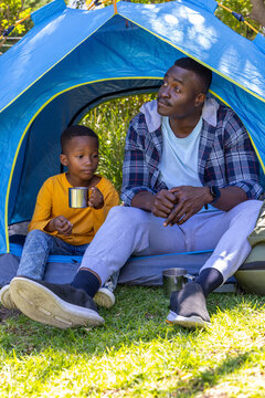 African American father and son sitting at tent entrance holding metal mugs, backpack nearby