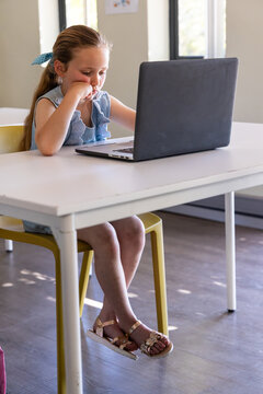 School-aged girl sitting at white table on yellow chair, wearing denim top, using laptop