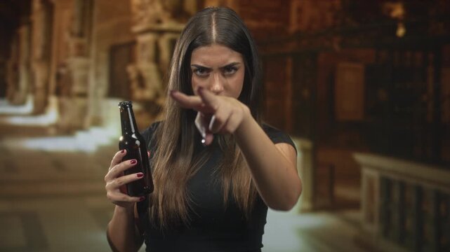 Woman holding beer bottle and making v sign over eyes while pointing finger toward camera in building; defiance confrontation.