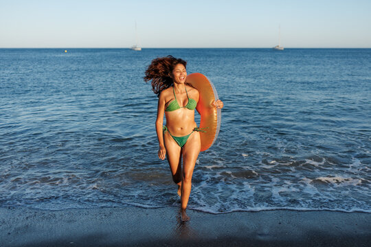 Woman in green bikini running from the ocean with an orange inflatable ring