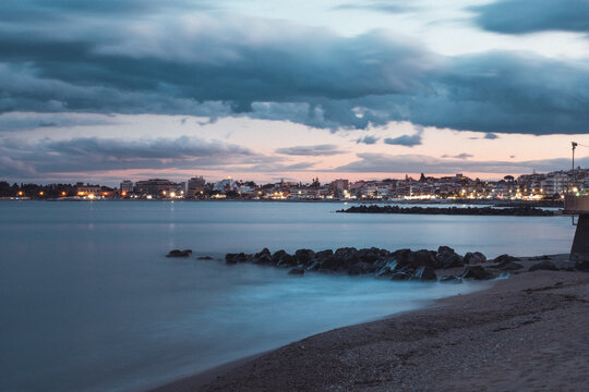 View of the tranquil beach with the city lights glowing softly under a dramatic sky with dark clouds, Taormina, Sicily, Italy.