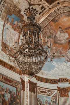 View of an ornate chandelier hangs beneath a frescoed ceiling depicting biblical scenes, creating a striking contrast of light and shadow, Taormina, Sicily, Italy.