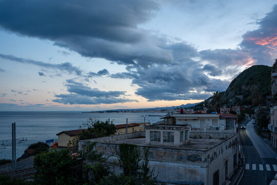 View of a dramatic coastal scene unfolds with the sea meeting the sky under a blanket of swirling clouds, a road winds beside buildings, Taormina, Catania, Italy.