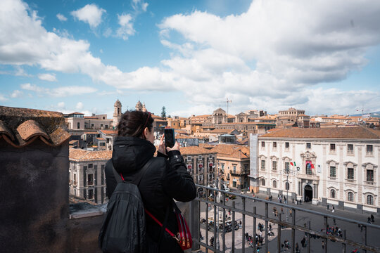 View of a woman capturing the warm hues of historic buildings and a bustling square under a bright, airy sky, Taormina, Sicily, Italy.