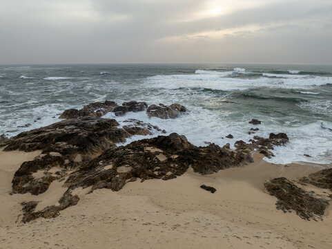 Aerial view of the turbulent ocean crashing against the rugged, dark rocks and sandy beach under a grey, overcast sky, Vila Cha, Portugal.