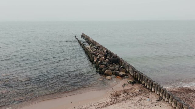 Wooden groynes and large rocks on the seashore. Coastal landscape with rough waves hitting barrier structures. Minimalist marine scenery for nature background.