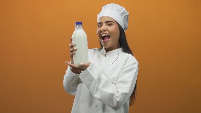 Woman chef holds milk bottle with both hands, mouth open in a playful kiss or singing gesture, studio setting with orange backdrop; dairy pride joy.