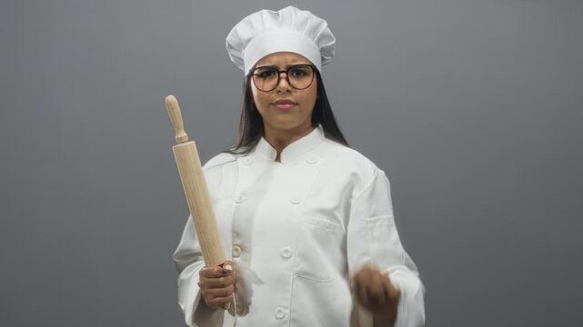Young hispanic woman chef in white coat and hat holds rolling pin and shows palm stop gesture in studio; determination safety.
