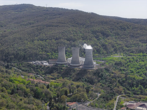 Larderello, Italy - 10 April 2026: Aerial view of the Enel Green Power geothermal power plant with three cooling towers emitting steam surrounded by lush green hills and forests.