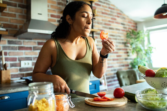 Young woman tasting fresh tomato while preparing salad in home kitchen