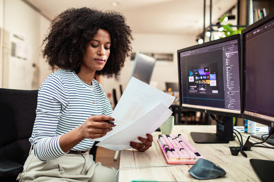 Female graphic designer reviewing documents at dual monitor workstation in modern office