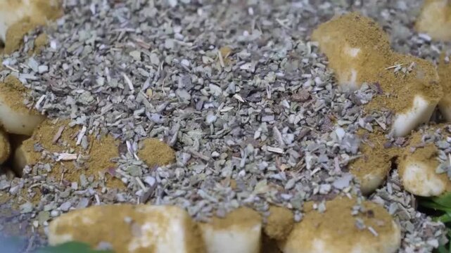 A high-angle, close-up shot of a hand sprinkling a mix of brown spices and greyish-green dried herbs onto chunks of light-colored food, possibly potatoes or chicken, during the cooking process. Focus 