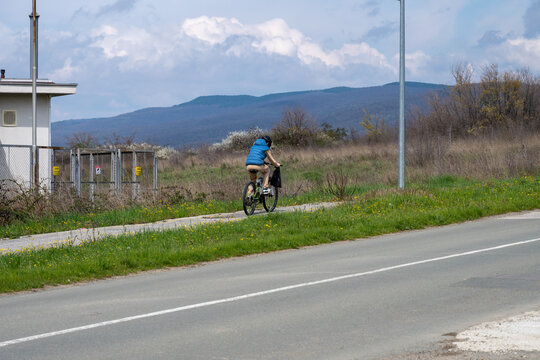 a person in a blue vest riding a bicycle along a path next to a paved road, with rolling hills and a cloudy sky in the bulgarian countryside.