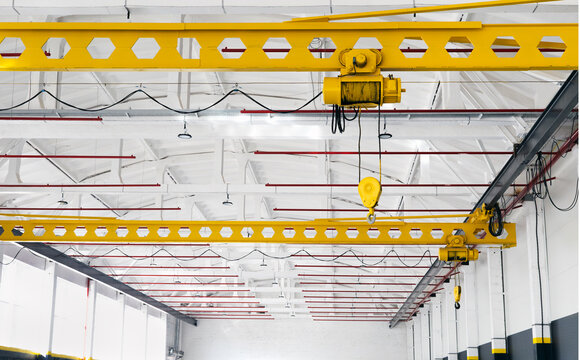 Yellow overhead crane with hook in a bright industrial hall. Steel beams and clean factory interior with visible red fire sprinkler system, heavy lifting equipment for manufacturing and logistics