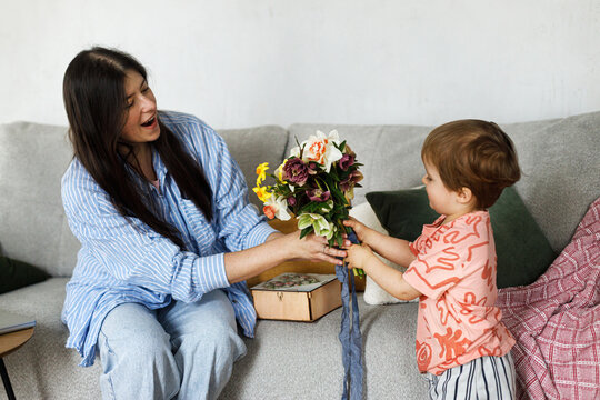 Happy Mothers day! Cute toddler son greeting mom and giving beautiful flowers, congratulation on mothers day. Stylish Mom and son together at home. Family domestic moments
