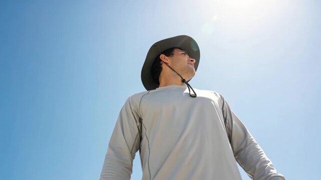 Young man wearing sun hat and long sleeve UV shirt looking up outdoors under bright sunlight, sun protection and outdoor adventure concept