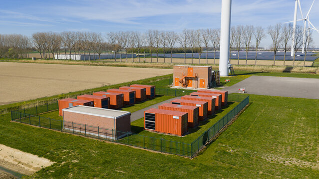 Aerial view of a battery energy storage system with orange containers and a brick building near wind turbines in Middelharnis, Zuid-Holland, Netherlands.