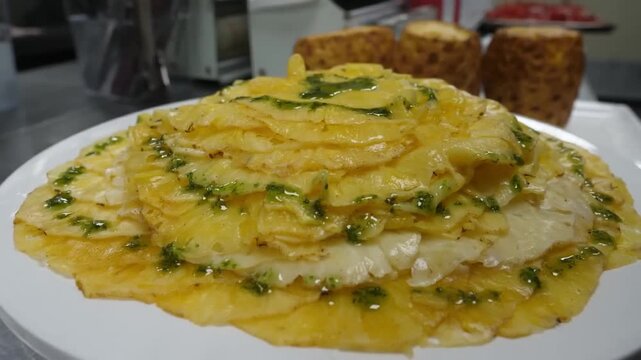 A close-up, top-down shot of a beautifully layered pineapple carpaccio, featuring thinly sliced pineapple arranged in a circular, overlapping pattern on a white plate. In the background, whole, cored 